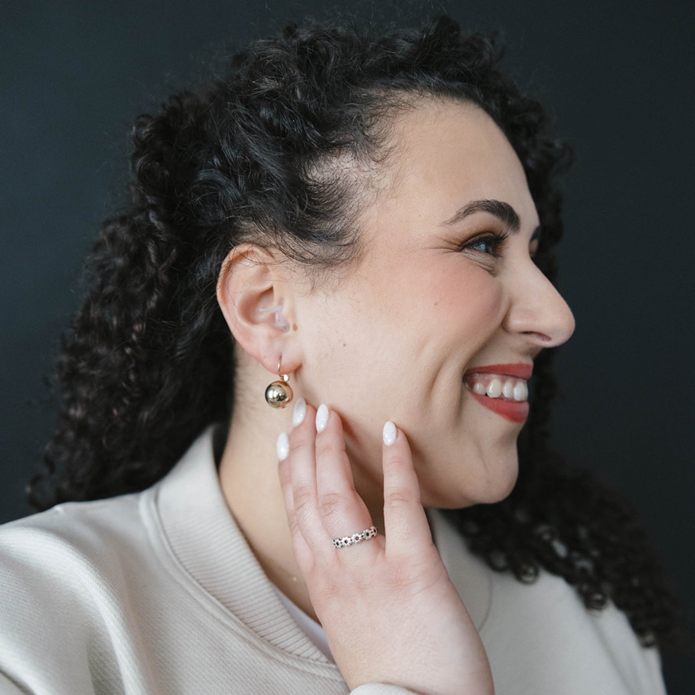 Woman wearing gold earrings and a ring, smiling against a dark background